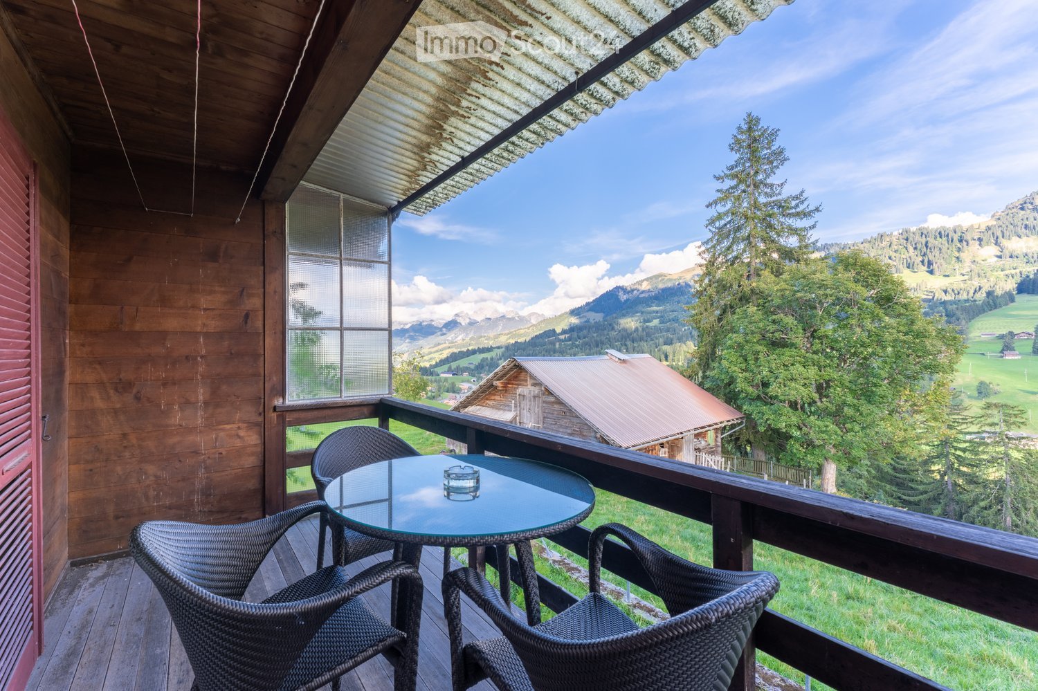 Wooden terrace, glass railing, round table, four wicker chairs, wooden flooring, wooden exterior wall, glass window, metal roof, scenic mountainous landscape, distant house, greenery, clear blue sky.