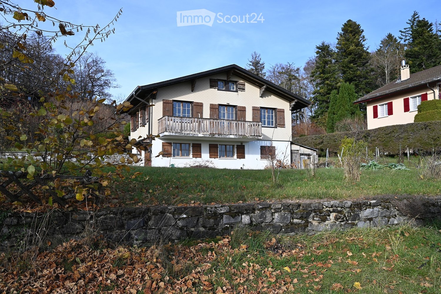 Two-story house with a balcony, wooden shutters, and a stone retaining wall in front.