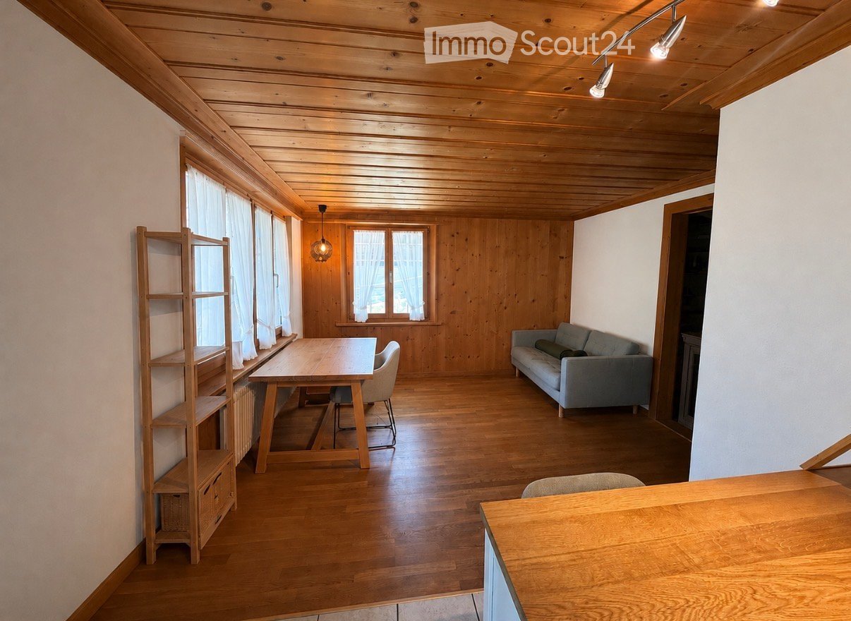 Bright living room with wooden walls and ceiling, large windows with white curtains, a grey sofa, a wooden dining table with chairs, and a wooden shelf.