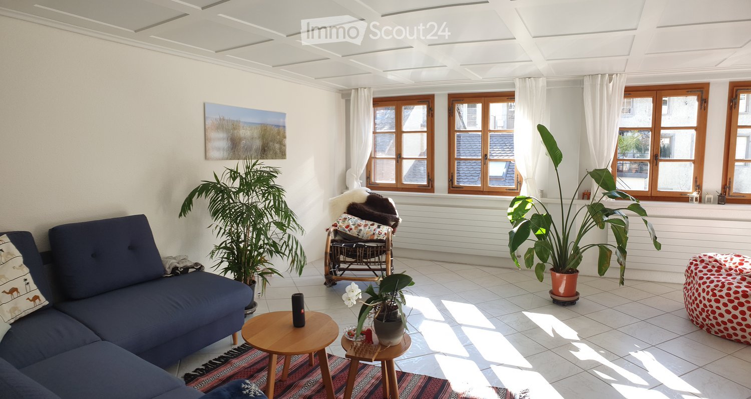 Bright living room with white walls, large windows, and a blue sectional sofa. Features include a wooden coffee table, a small wooden stool, potted plants, a decorative chair, and a picture frame on the wall.