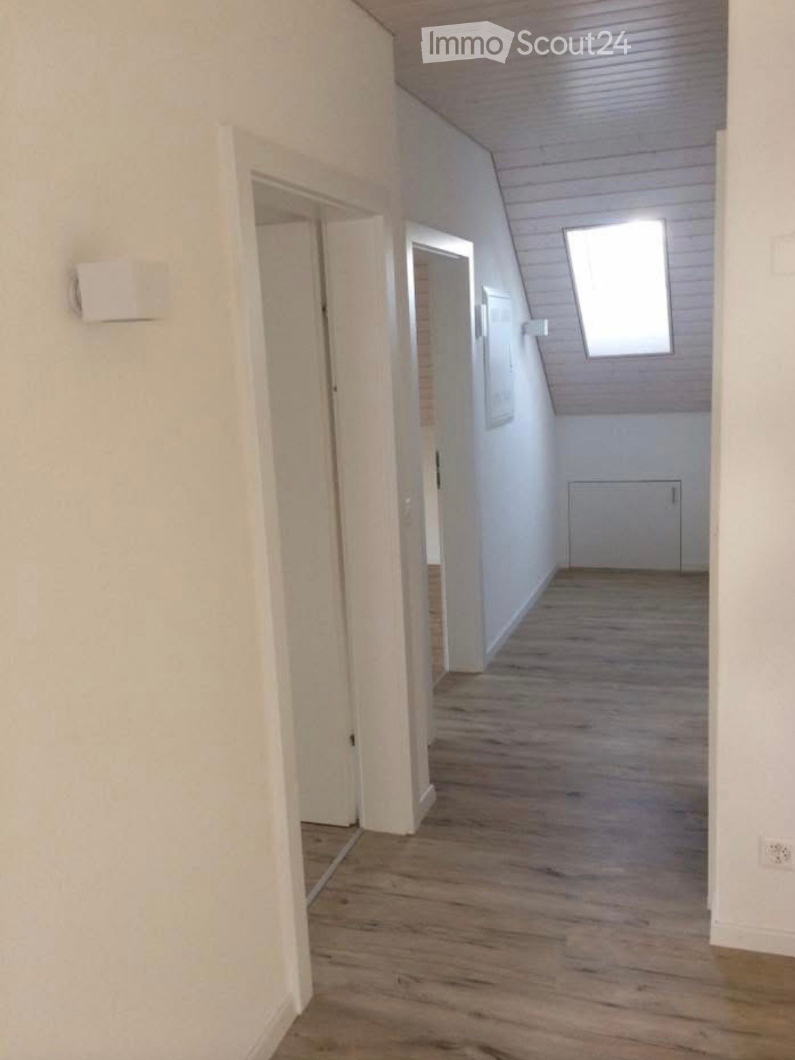 Empty hallway with wooden flooring, white walls, and multiple doors leading to other rooms. A small window is visible at the far end.