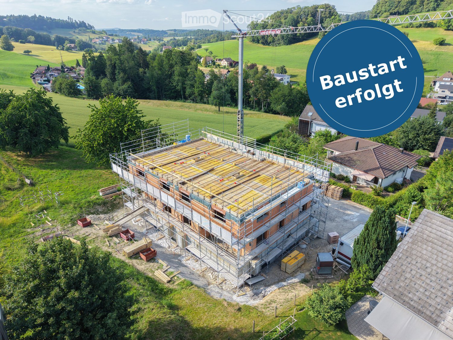 Aerial view of a residential construction site with scaffolding, exposed wooden beams, and a crane in the background. The surrounding area includes green fields, trees, and neighboring houses.