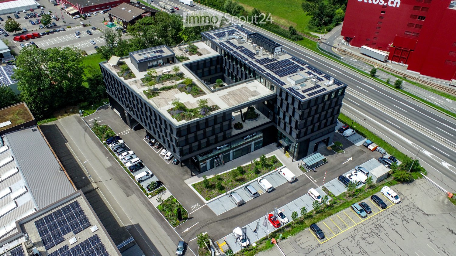 Modern multi-story building with solar panels on the roof, surrounded by a parking lot with numerous cars and a nearby road.