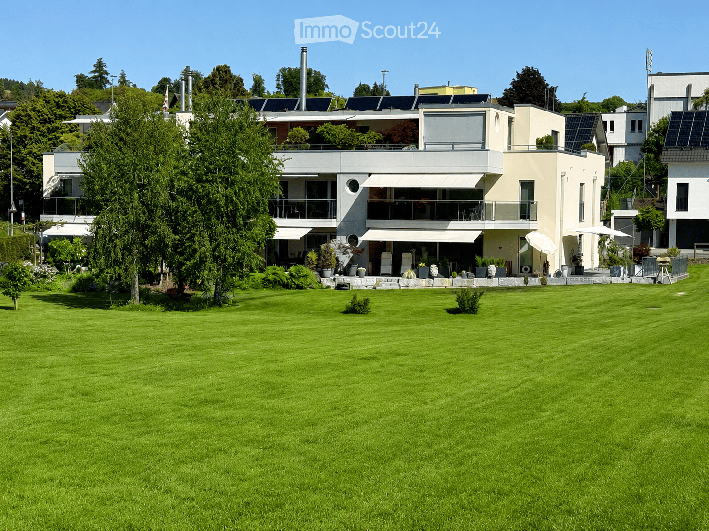 Modern two-story house with large windows, balconies, and solar panels on the roof, surrounded by a well-maintained lawn and trees.