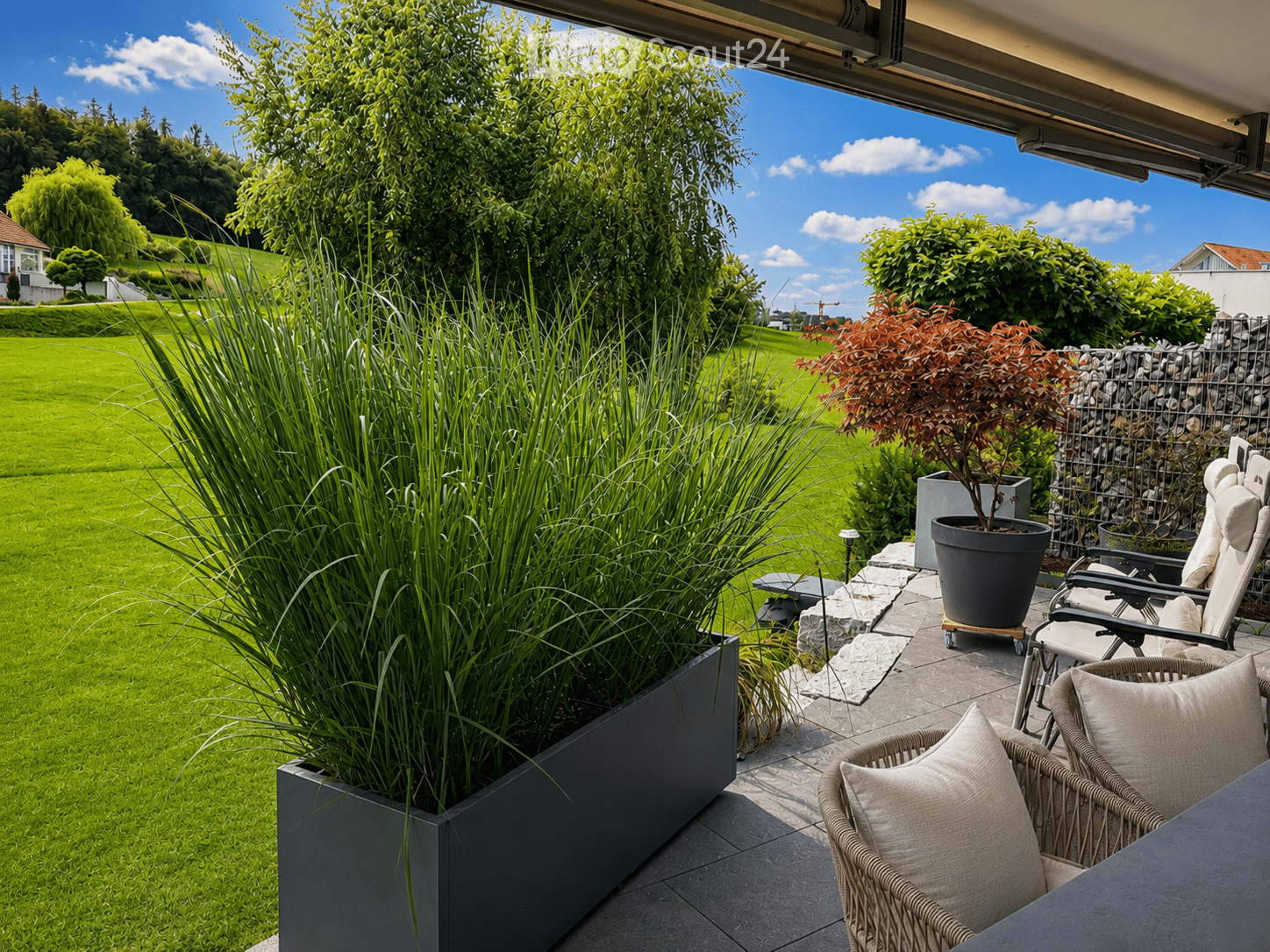 A well-maintained garden with a patio area featuring wicker chairs, a table, and potted plants, overlooking a lush green lawn and trees.