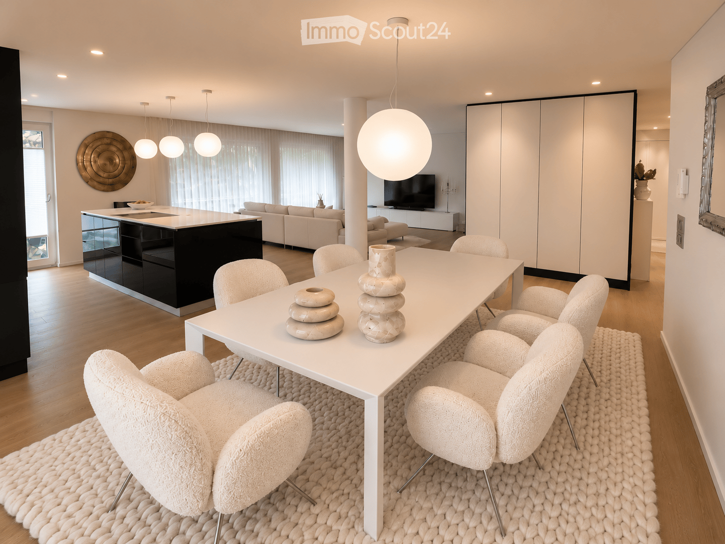 Modern, open-plan living and dining area with a white dining table, six white chairs, a black kitchen island, a light grey sofa, a flat-screen TV, decorative vases, and pendant lights.