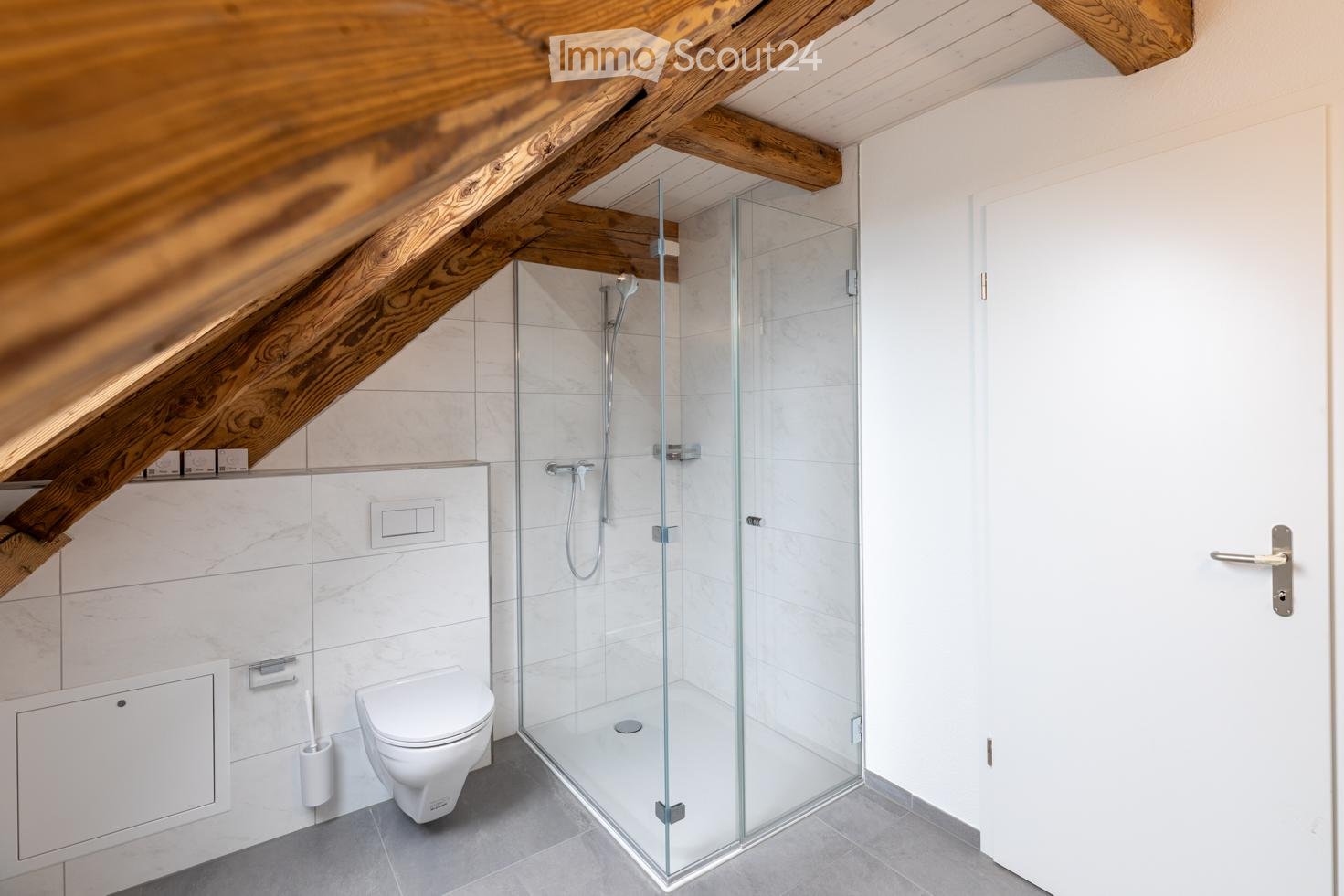 Modern bathroom with white tiles, a glass-enclosed shower, toilet, and wooden slanted ceiling beams.