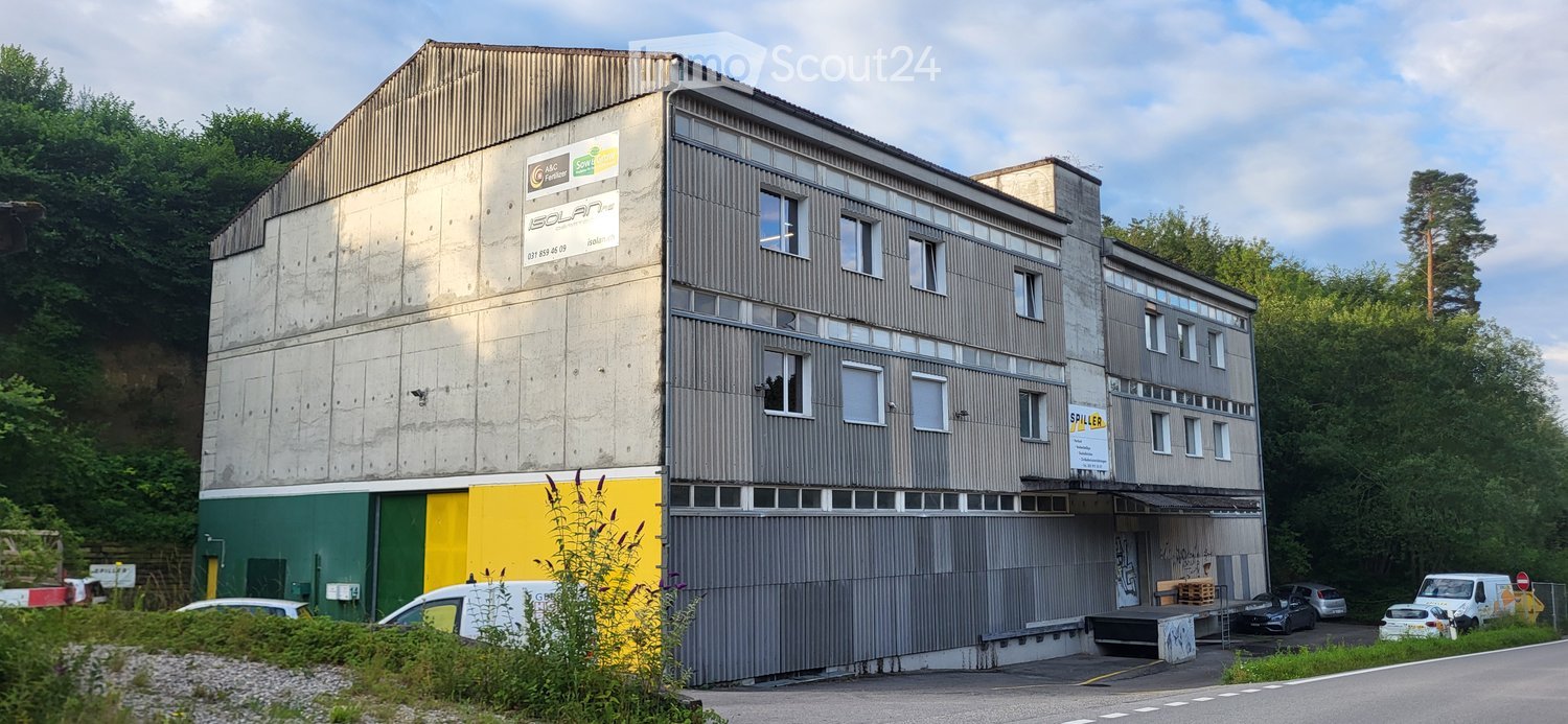 Multi-story industrial building with a mix of concrete and metal siding, featuring signage for 'Solana' and 'Spiller', surrounded by greenery and parked vehicles.