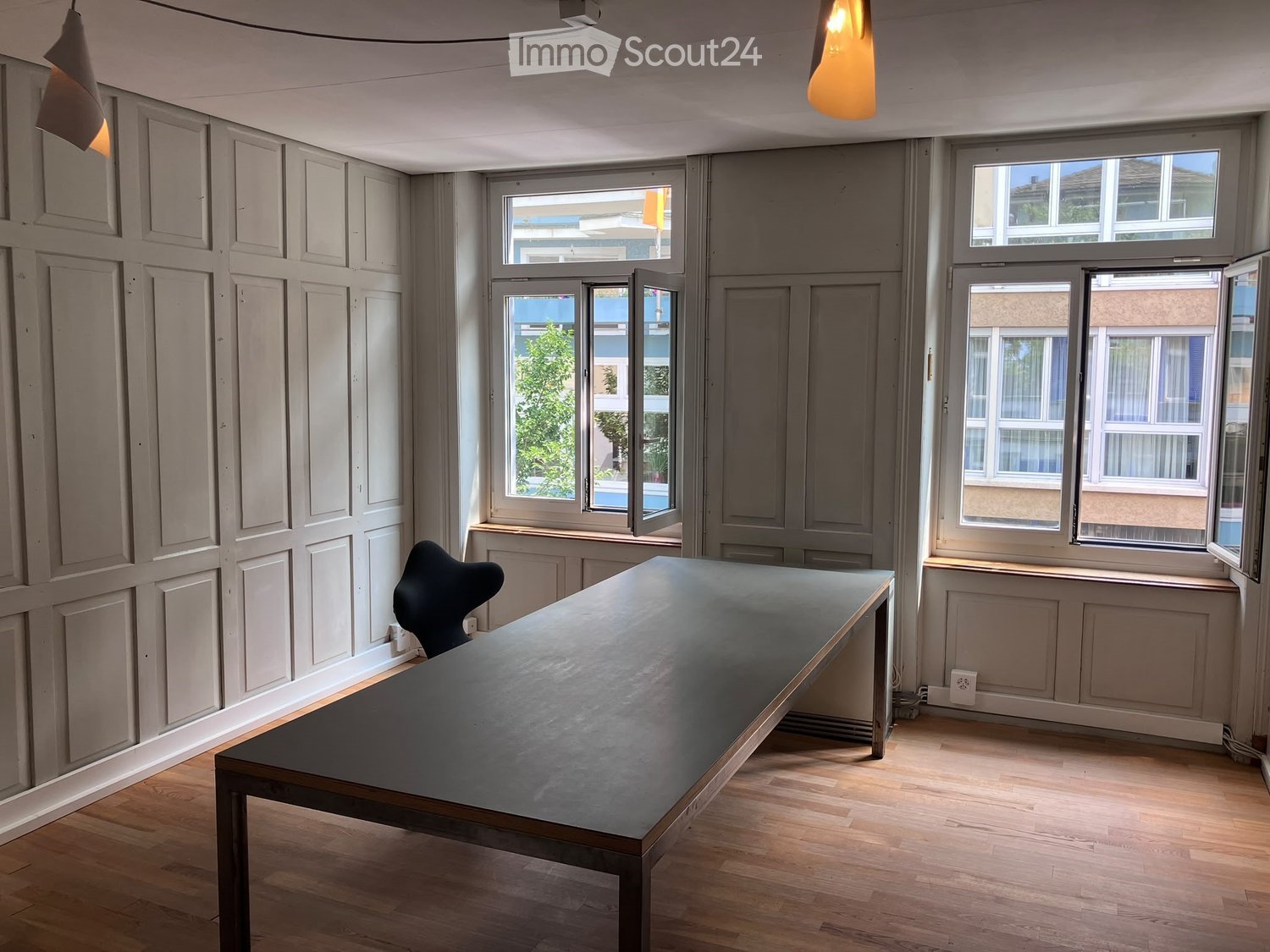 Minimalist room with wooden flooring, grey walls, large windows, a long dark table, and a single chair.