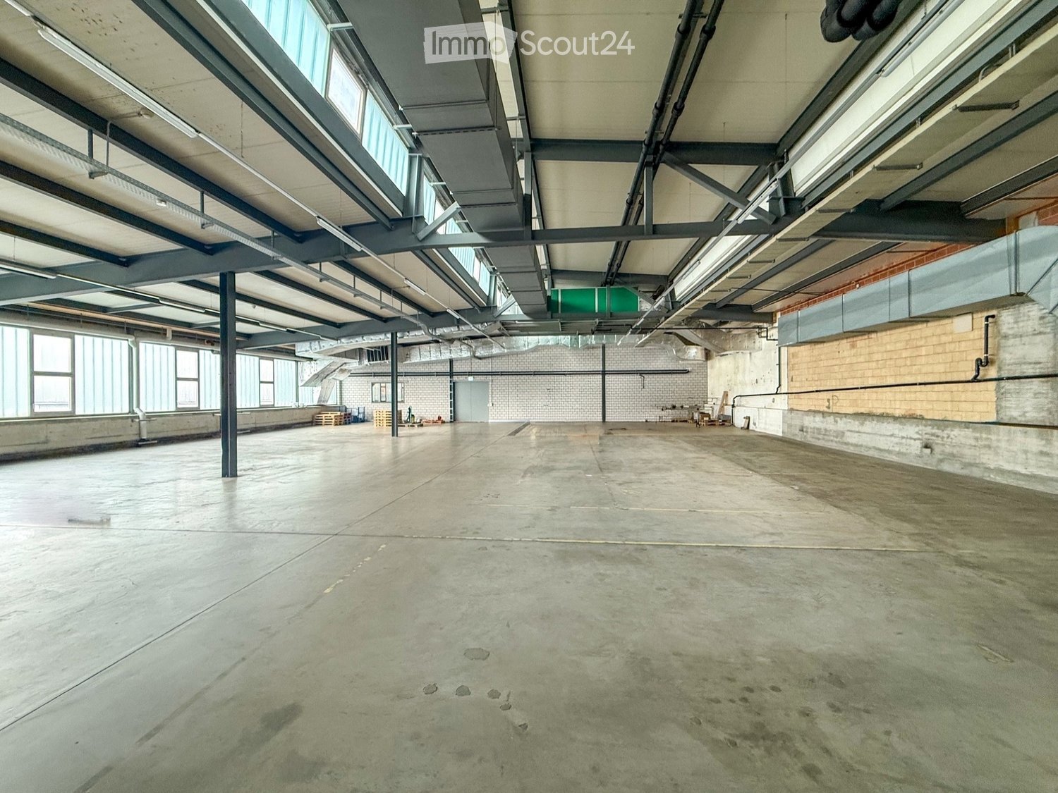 Large, empty industrial-style space with concrete floors, exposed metal beams, and large windows allowing natural light. Minimal furnishings, including a few stacked crates and a small table in the background.