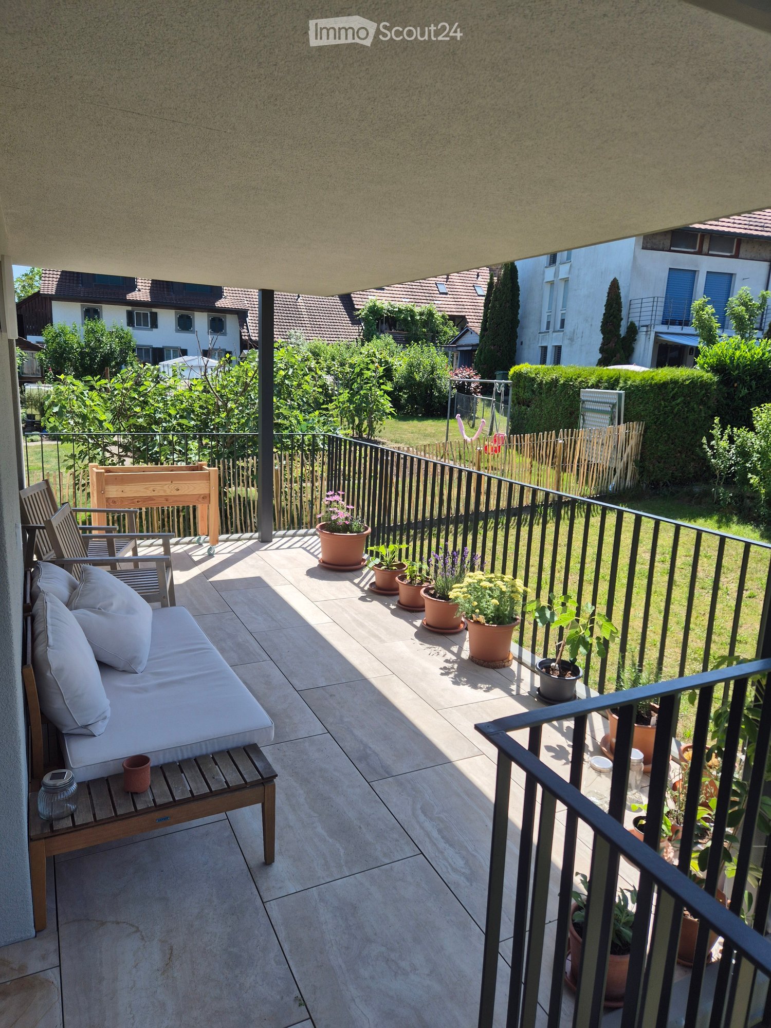 Balcony with a sofa, chairs, a small table, and several potted plants, overlooking a garden area with trees and houses.