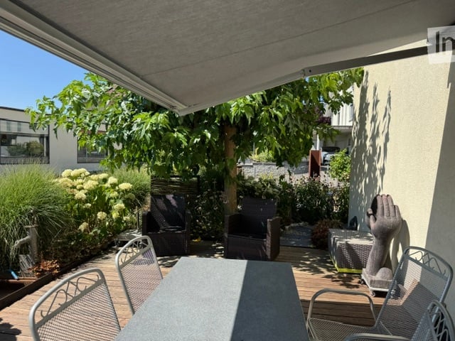 A covered outdoor terrace with wooden decking, surrounded by greenery and plants. There are several chairs and a table, with a large tree providing shade.