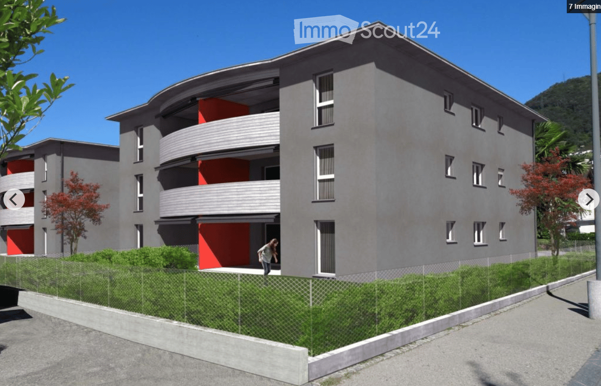 Modern multi-story residential building with grey facade, red accents, balconies, and surrounding greenery.