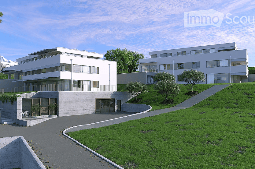 Modern multi-story residential building with clean lines, large windows, and a garage entrance. The structure is set on a raised ground floor with a landscaped green area and pathway leading up to the building.