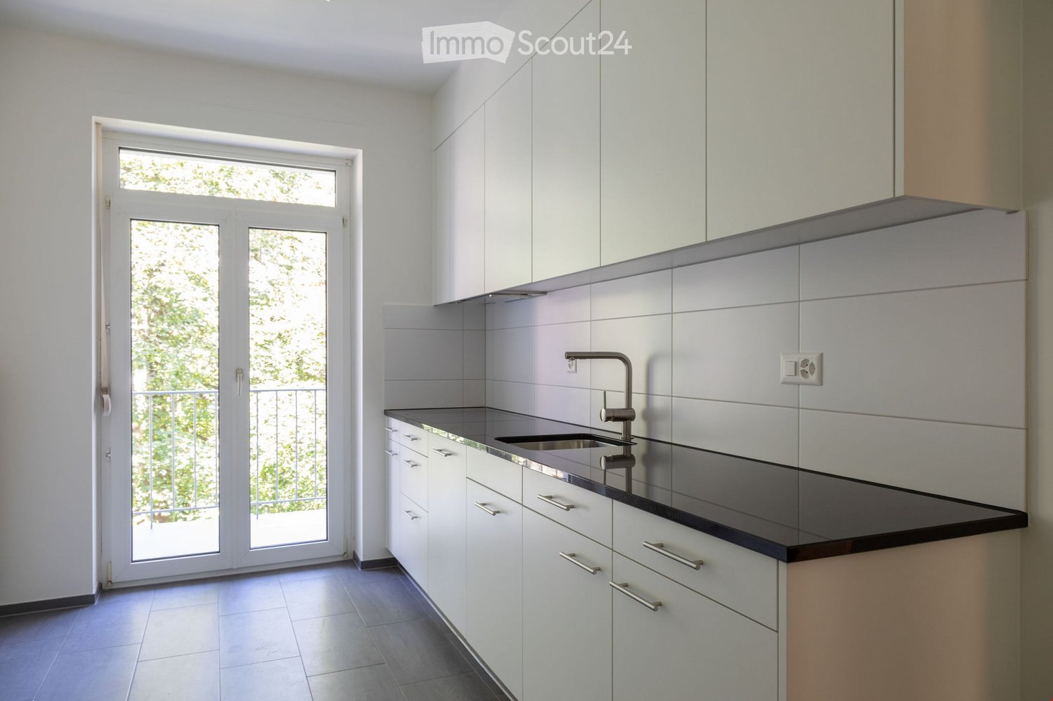 Modern kitchen with white cabinets, dark countertop, and a sink with a modern faucet. A large window and sliding glass door lead to a balcony.