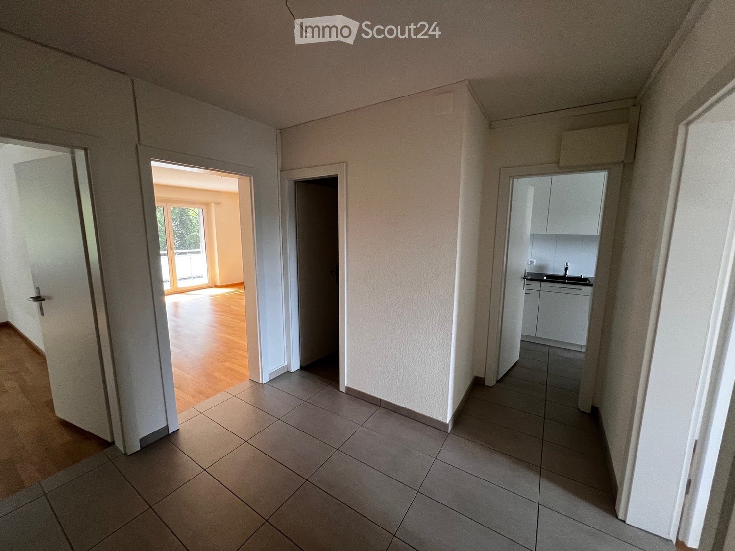 Empty hallway with tiled floor, white walls, and doors leading to other rooms, including a kitchen area with white cabinets and a sink.
