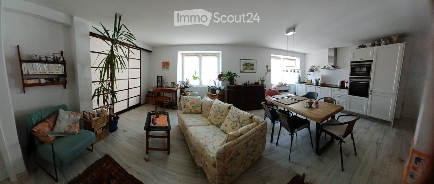 A well-lit living and dining area with a floral-patterned sofa, coffee table, dining table with chairs, and a kitchen area with white cabinets and appliances.
