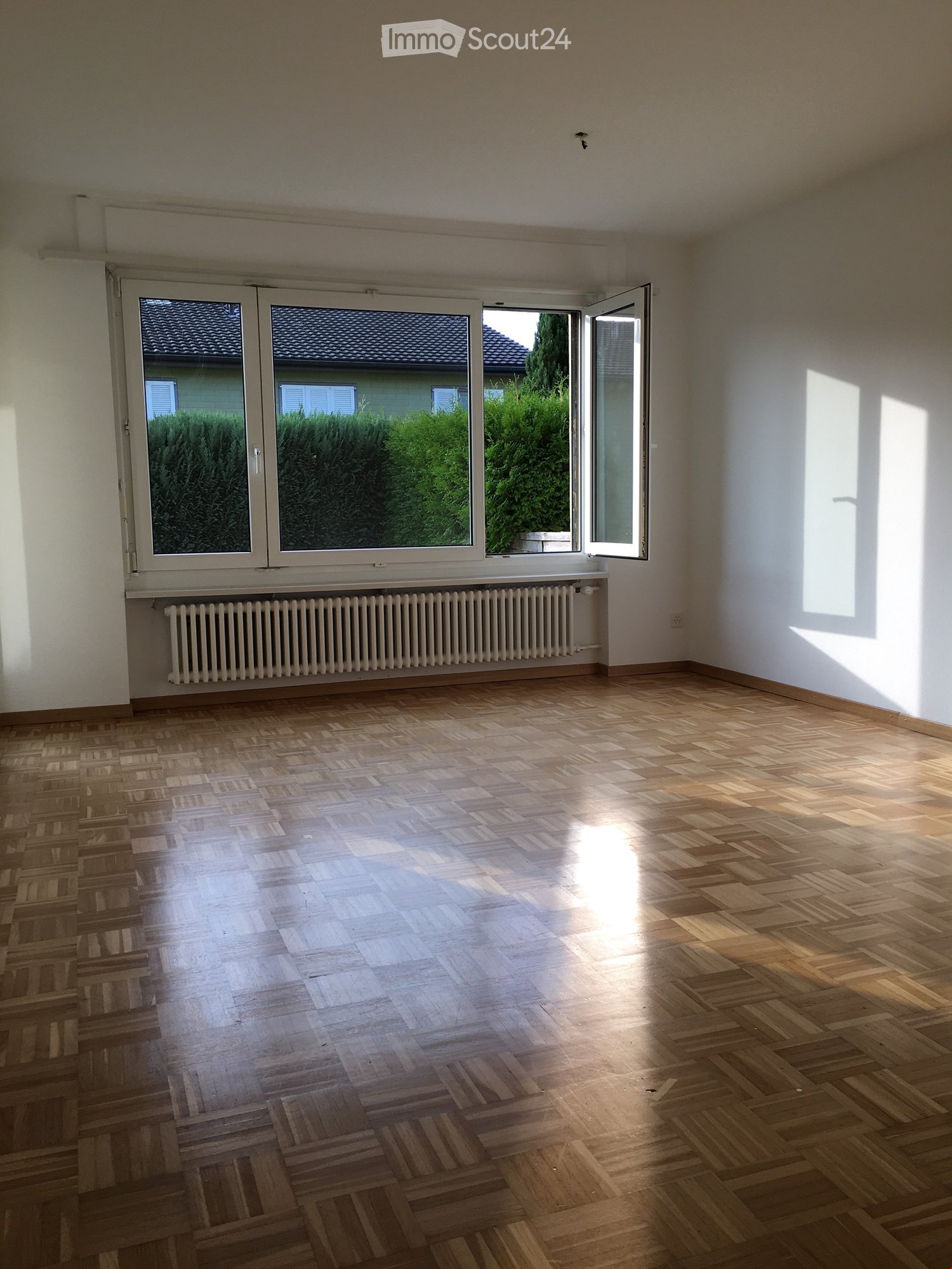 Empty room with wooden parquet flooring, white walls, large windows with white frames, and a radiator below the windows.