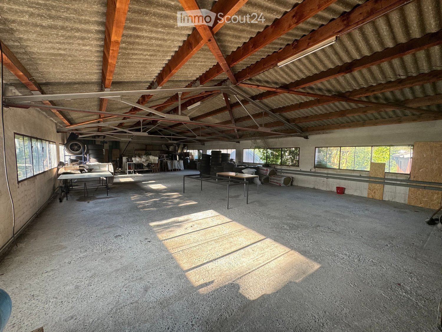 An empty, spacious room with a concrete floor and exposed wooden beams on the ceiling. The room has large windows allowing natural light to enter, and there are some storage items and equipment visible in the background.