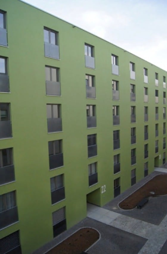 Modern multi-story building with green facade and numerous windows, featuring a numbered entrance and small landscaped areas in front.