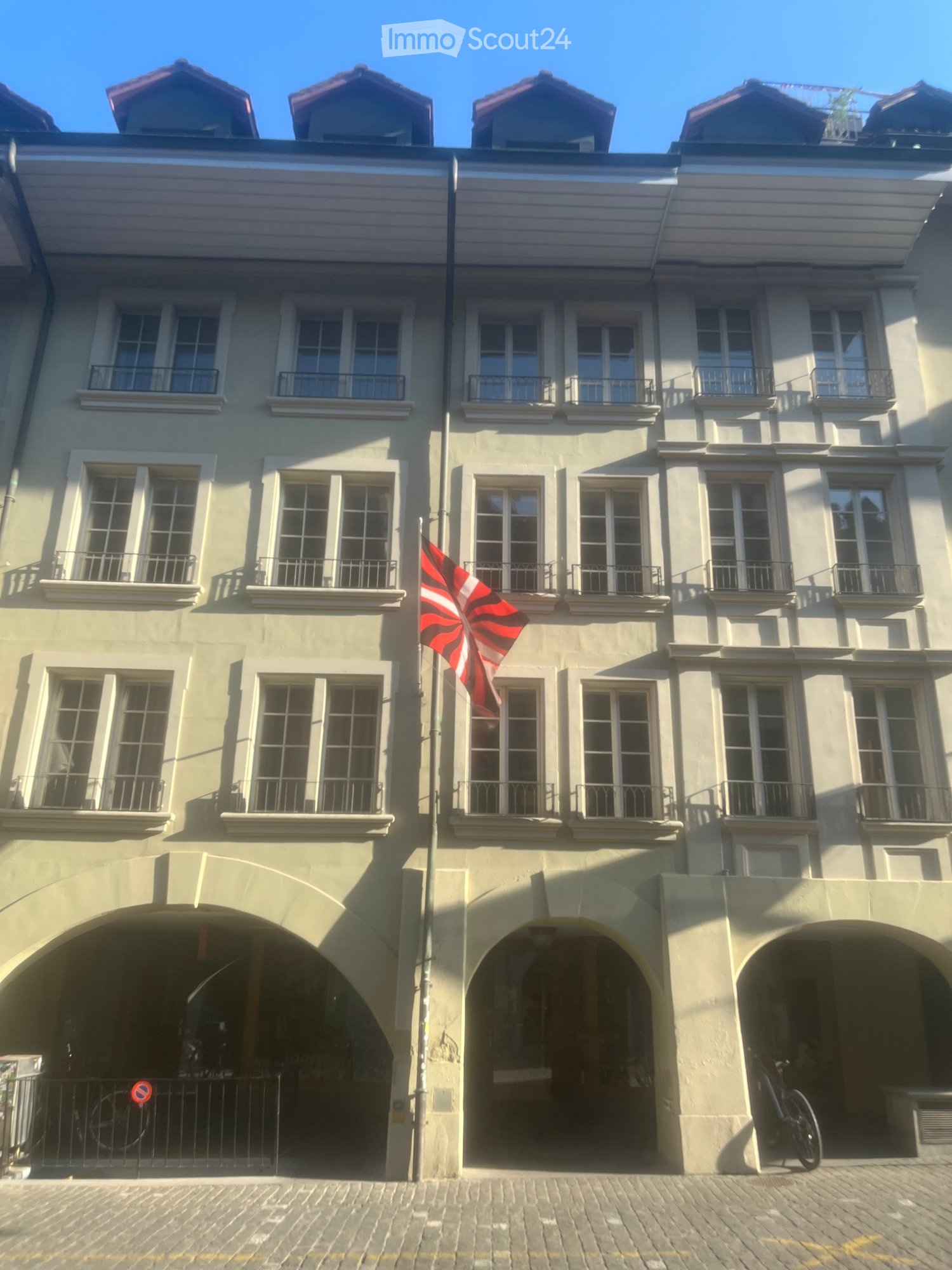 A multi-story building with light yellow facade, multiple windows with white frames, and balconies. A red and white flag is displayed on a pole in front of the building. There are three arched entrances at the bottom, and a bicycle is parked near the righ
