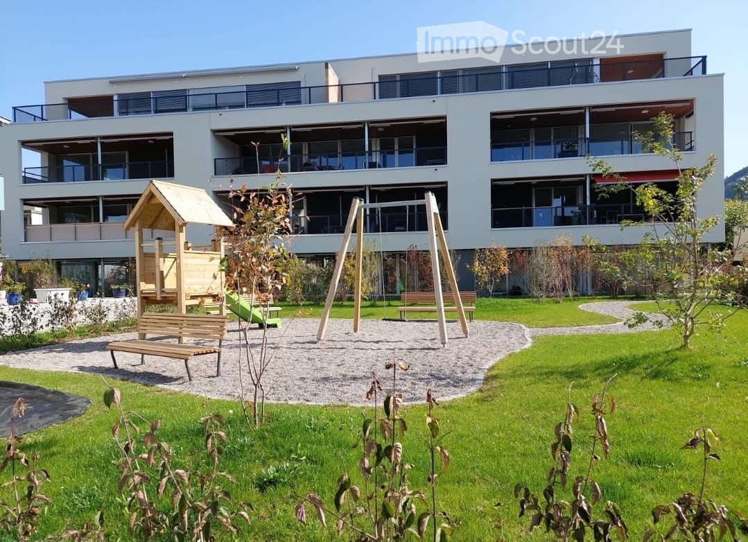 Modern multi-story building with balconies, surrounded by a well-maintained garden area featuring a playground with swings, benches, and a small wooden playhouse.