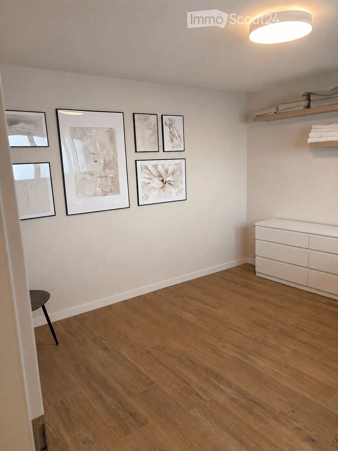 Empty room with wooden flooring, white walls, and modern decor including framed artwork and a small cabinet with shelves.