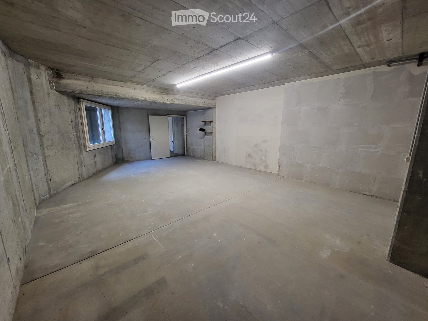 Empty concrete room with a single window, a door leading to another space, and a small shelf in the background.