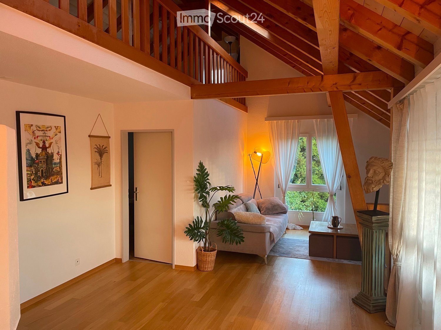 Bright living room with wooden floors, exposed wooden beams, large windows with white curtains, a sofa, a small table, a plant, and artwork on the walls.