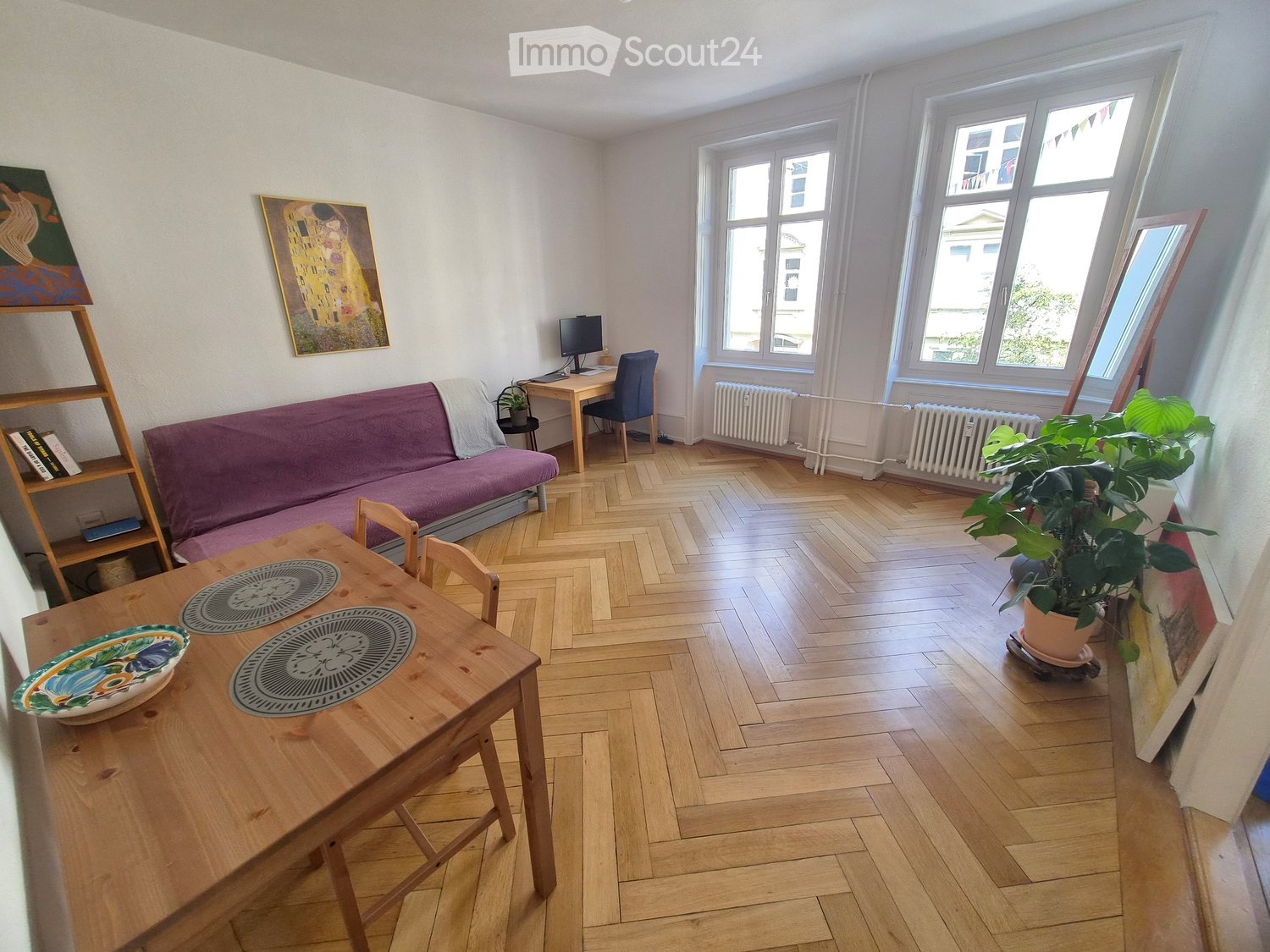A cozy living room with wooden flooring, a purple sofa, a wooden dining table with placemats and a decorative bowl, a small desk with a laptop, a potted plant, and artwork on the walls.