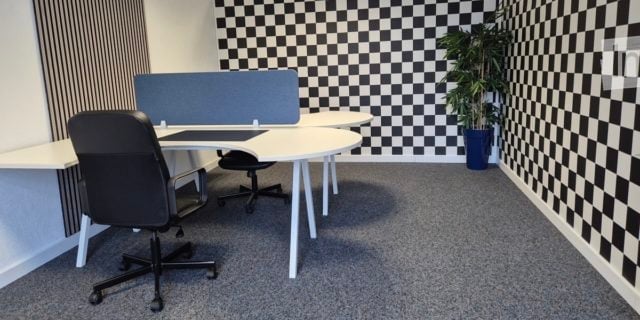 Modern office space with white desks, black office chairs, a computer screen, and a potted plant. The walls feature a black and white checkered pattern.