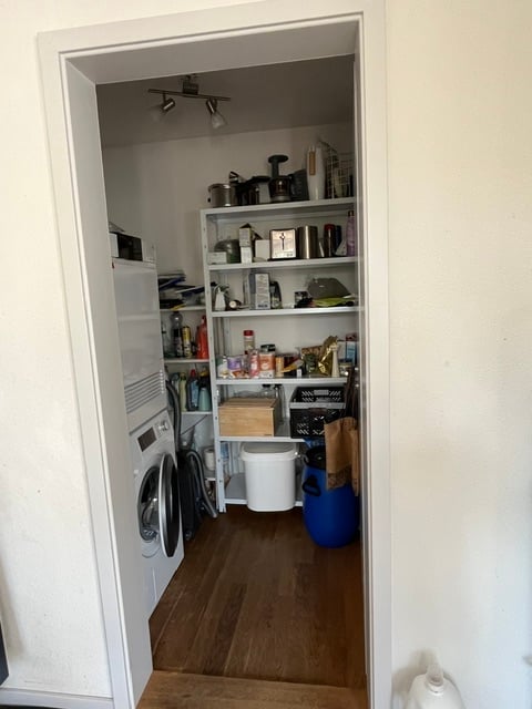 Open pantry area with wooden flooring, white walls, and shelves stocked with various kitchen items, including pots, pans, and boxes. A washing machine is visible to the left.