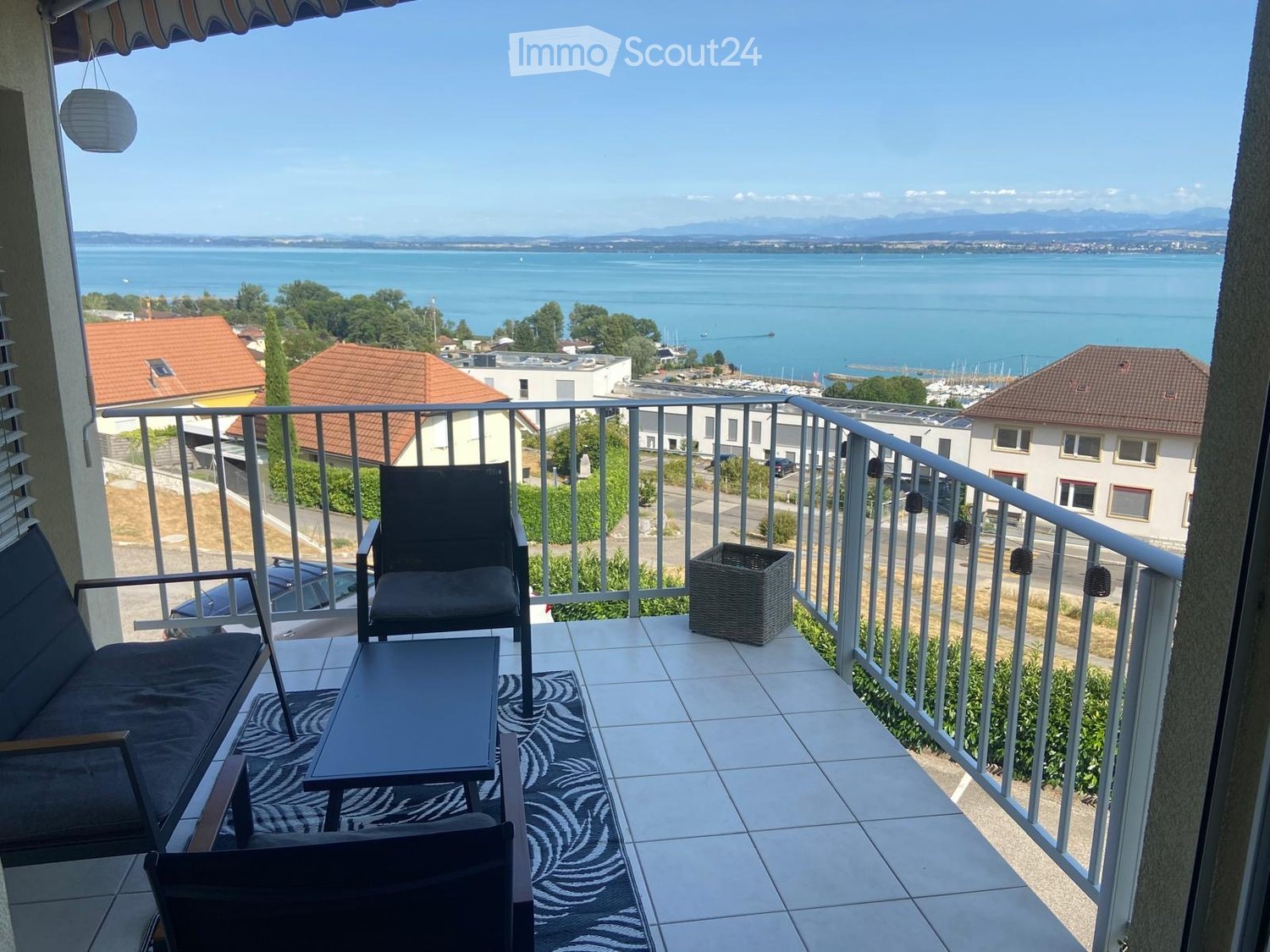 Balcony with a scenic view of a lake and distant mountains, furnished with a sofa, chairs, a table, and a wicker basket.