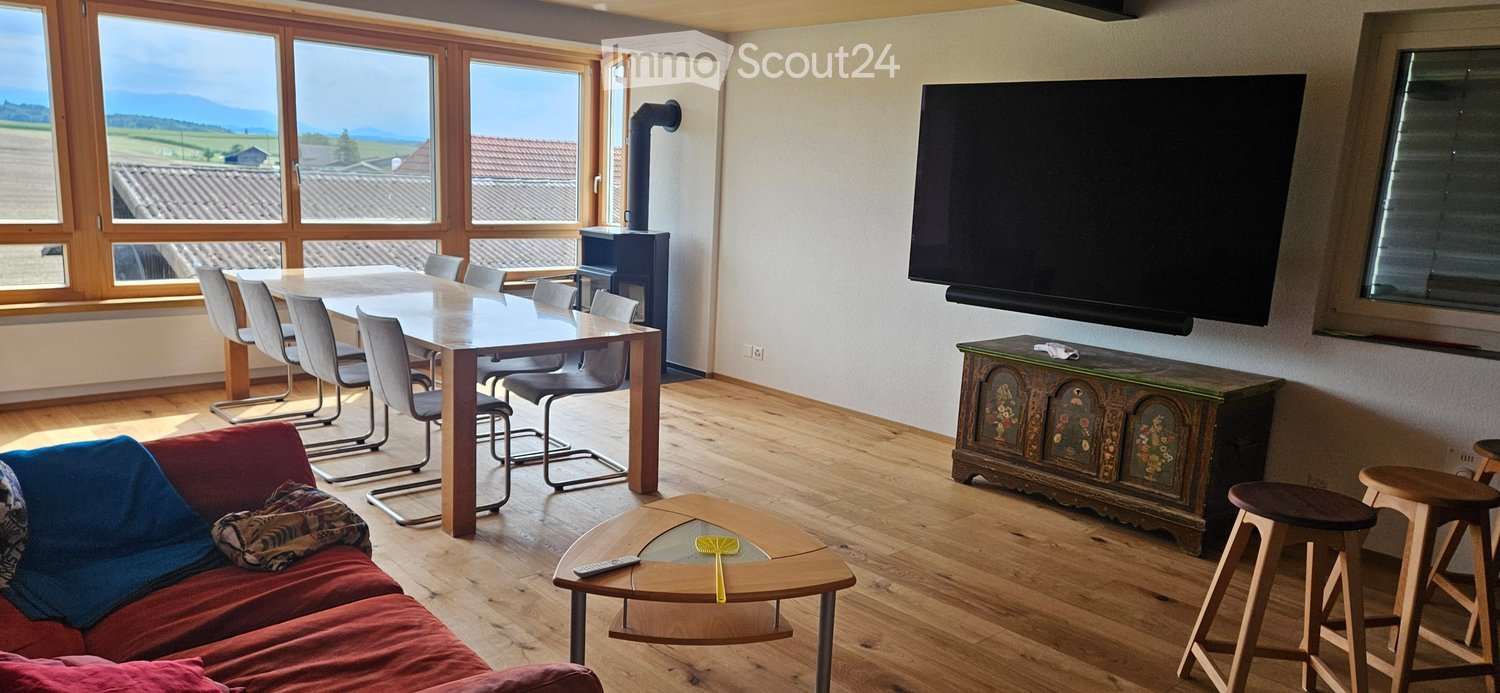 Bright living and dining area with wooden floors, large windows offering a scenic view, a red sofa, a coffee table, a dining table with chairs, and a fireplace.