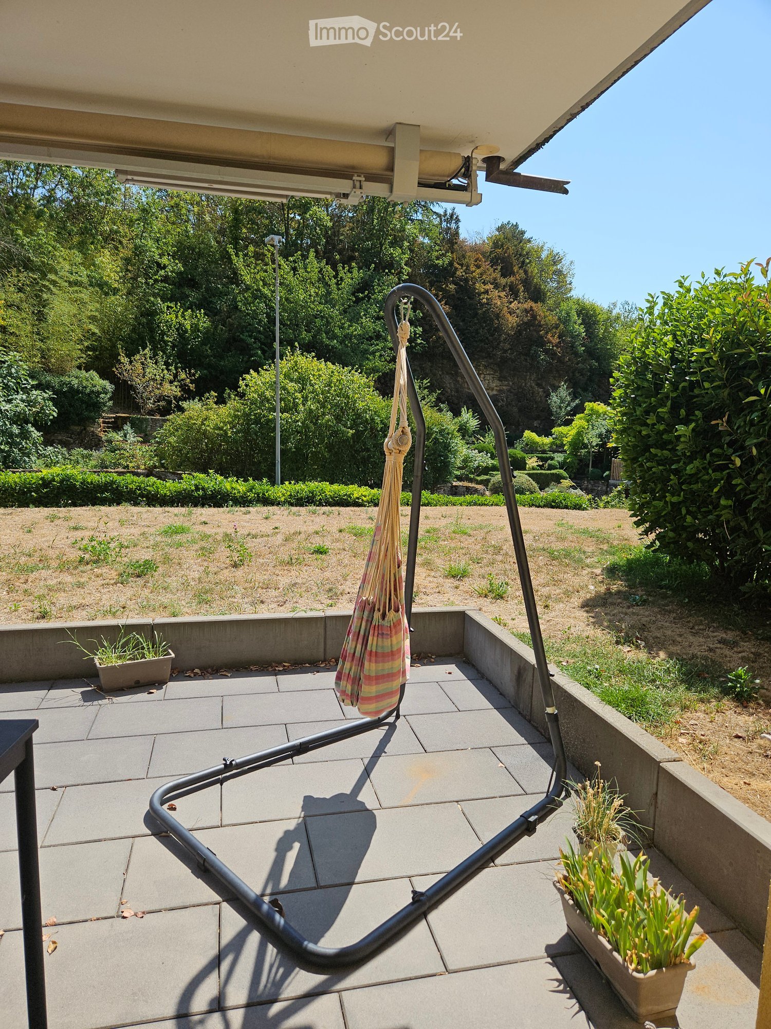 Balcony with tiled floor, metal railing, hanging striped bag, potted plants, and a view of a landscaped garden.