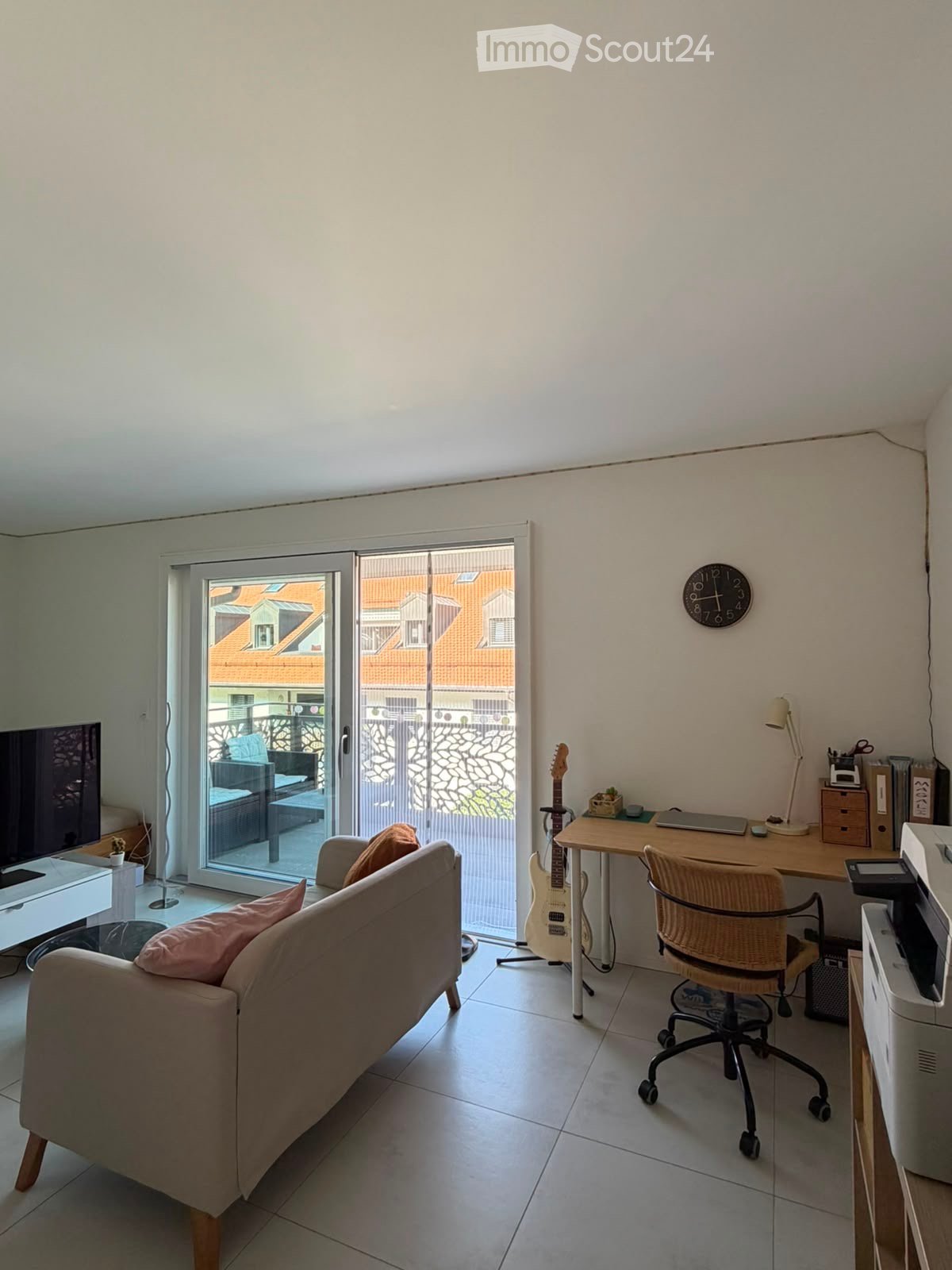 Bright living room with a sofa, desk, guitar, and sliding glass door leading to a balcony.