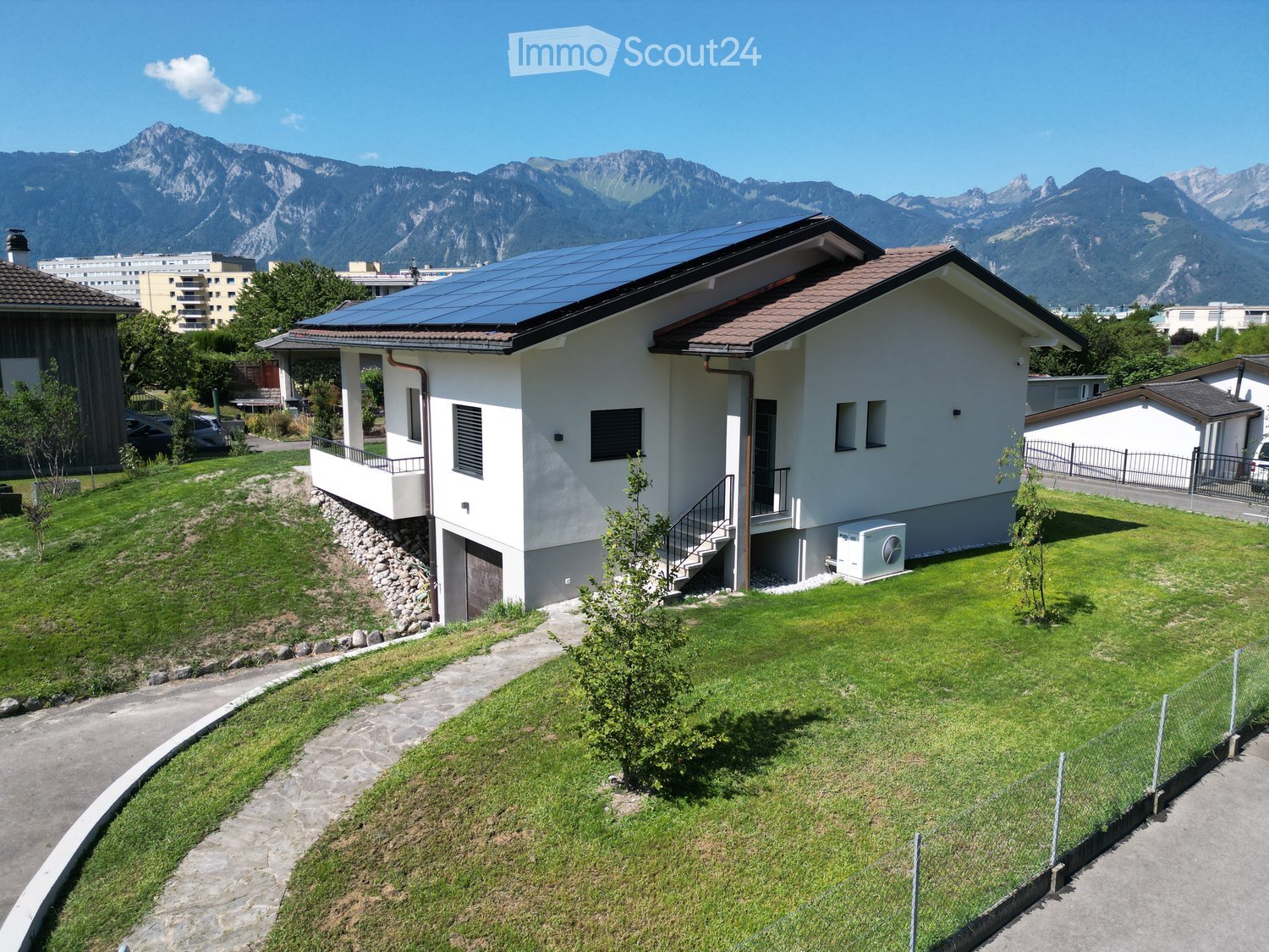 A modern two-story house with a solar panel roof, surrounded by a well-maintained lawn and small garden, with a scenic mountain backdrop.