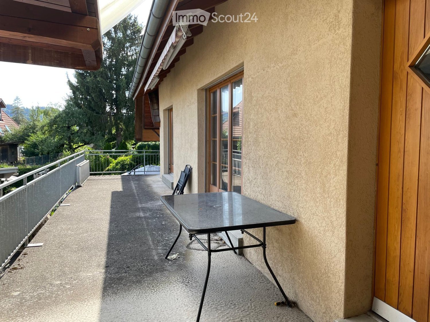 Balcony with a glass table and a single chair, overlooking a green area with trees and neighboring houses.