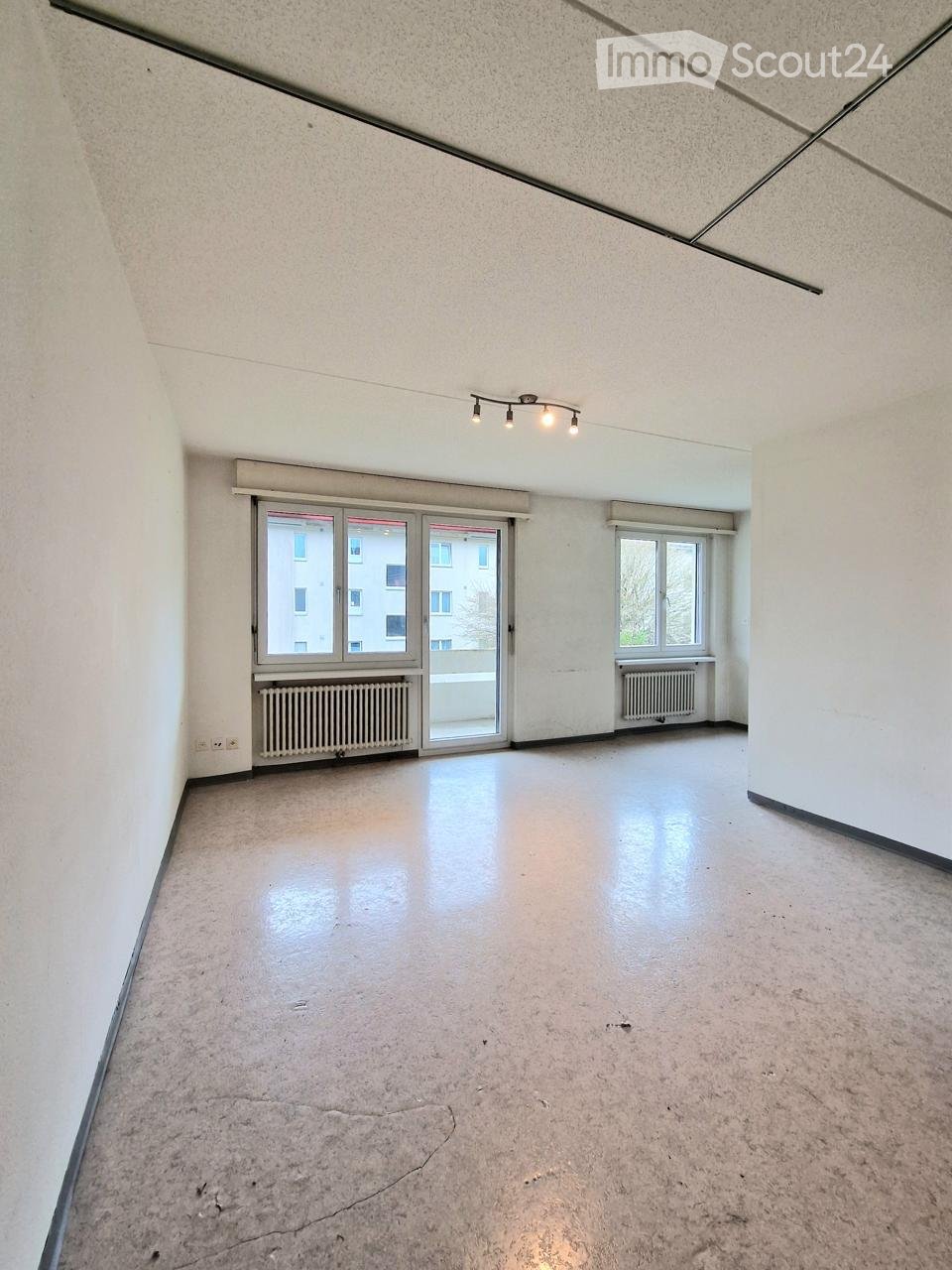 Empty room with white walls, concrete floor, large windows, radiators, and a sliding glass door leading to a balcony.