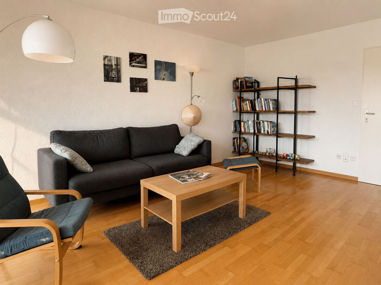 Modern living room with wooden flooring, grey sofa, wooden coffee table, armchair, bookshelf, and decorative picture frames on the wall.