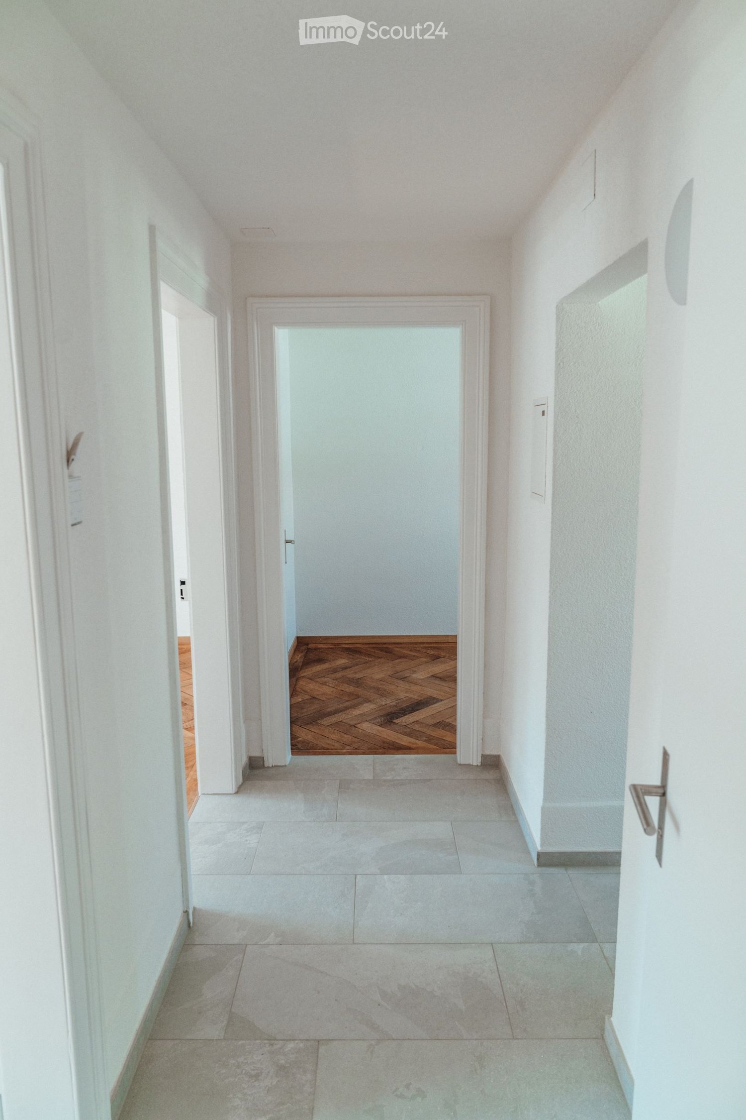 A clean, modern hallway with white walls, tiled flooring, and a wooden door at the end.