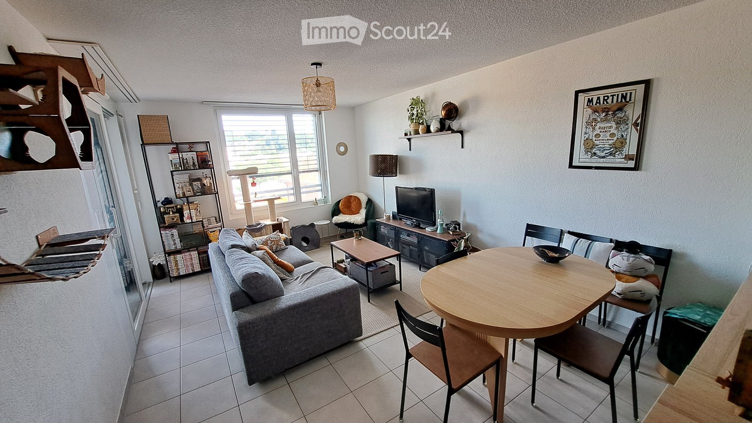 A modern living room with a grey sofa, coffee table, dining table with chairs, cat tree, shelves, and a television. The room features large windows and a cozy, well-lit atmosphere.
