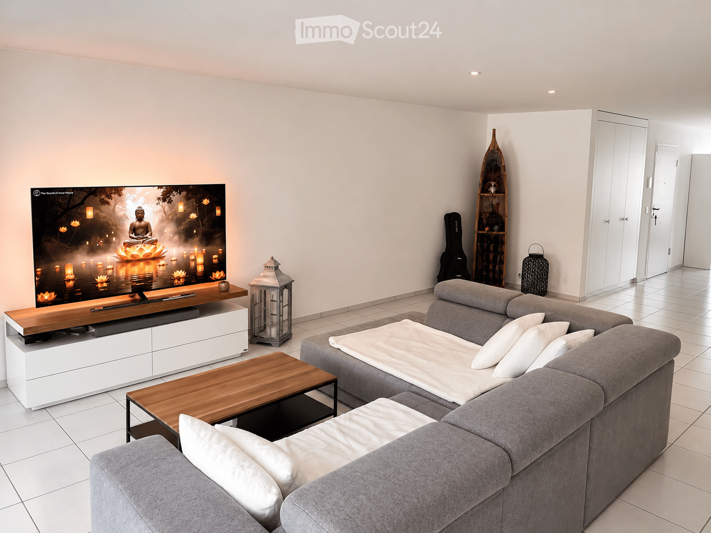 Modern living room with a grey sectional sofa, wooden coffee table, flat-screen TV on a white cabinet, decorative lantern, and a guitar case in the corner.