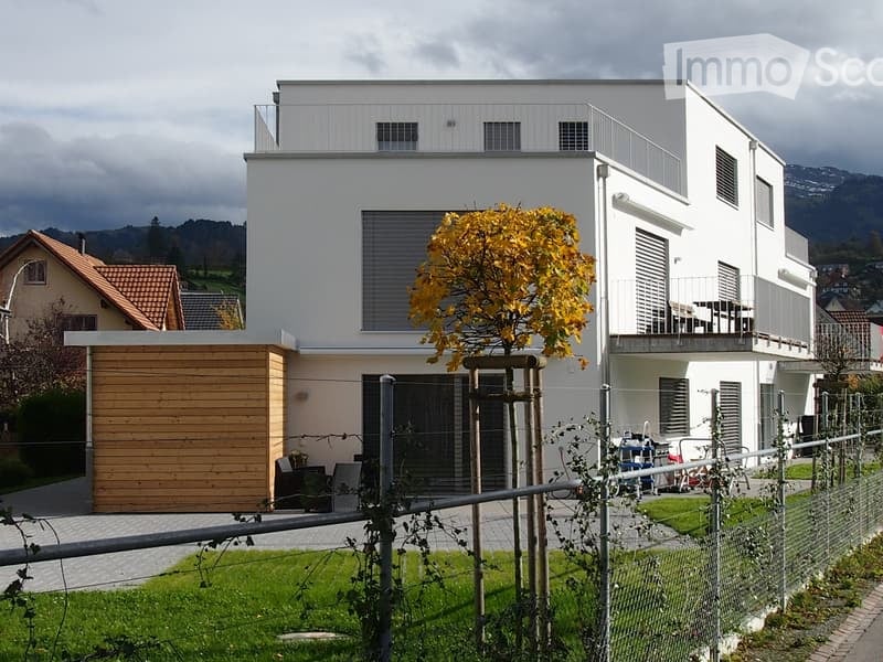 Modern two-story house with white exterior, large windows, and a balcony on the upper floor. Features a wooden garage and a small garden area with a tree.