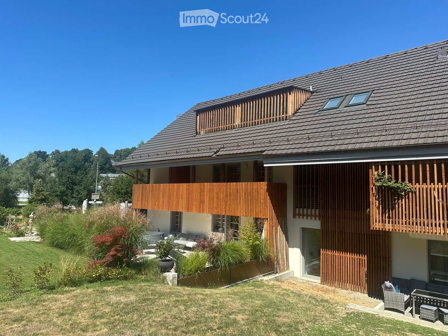Modern two-story house with wooden balconies, large windows, and a well-maintained garden with seating area.