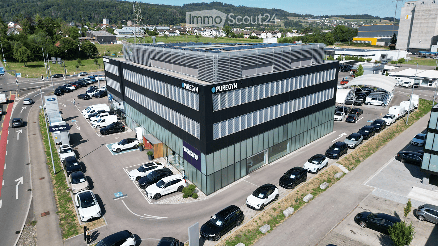 Modern commercial building with glass facade, labeled "PUREGYM" and "VOLVO" on the side, surrounded by a parking lot with numerous cars.