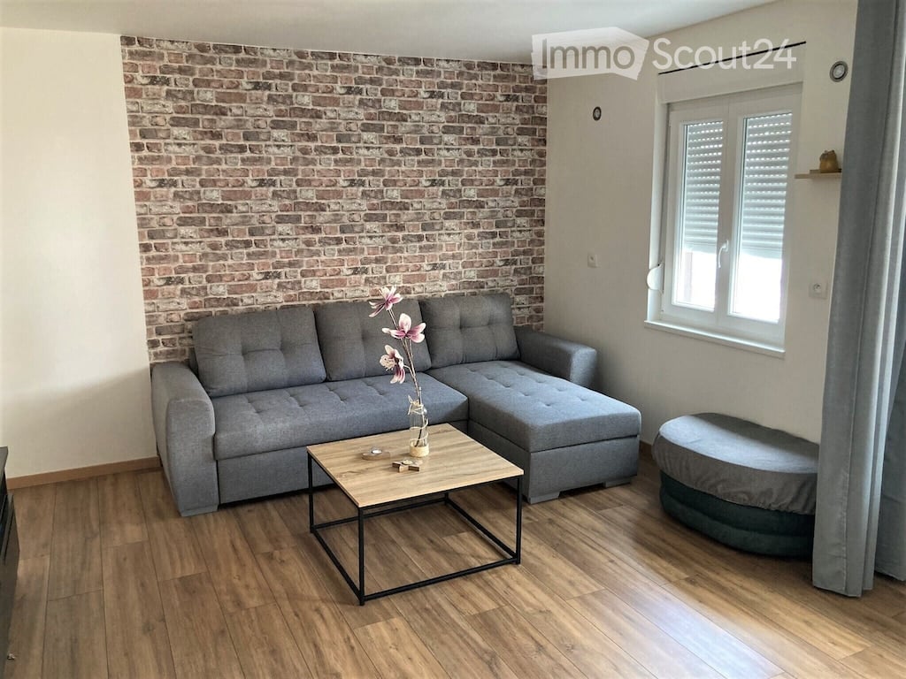 Living room with a grey sectional sofa, wooden coffee table, ottoman, and a vase with flowers on the coffee table. The room features a brick accent wall and wooden flooring.