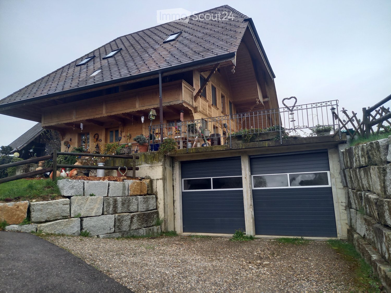Two-story house with a raised ground floor, wooden balcony, garage, and stone retaining wall.