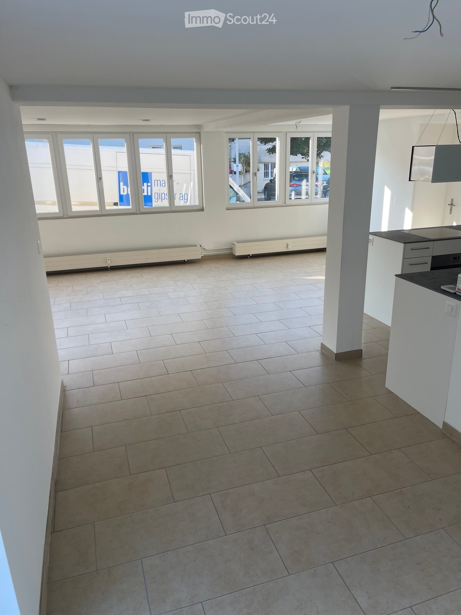 Empty room with tiled floor, white walls, large windows, and a small kitchen area with cabinets and a sink.