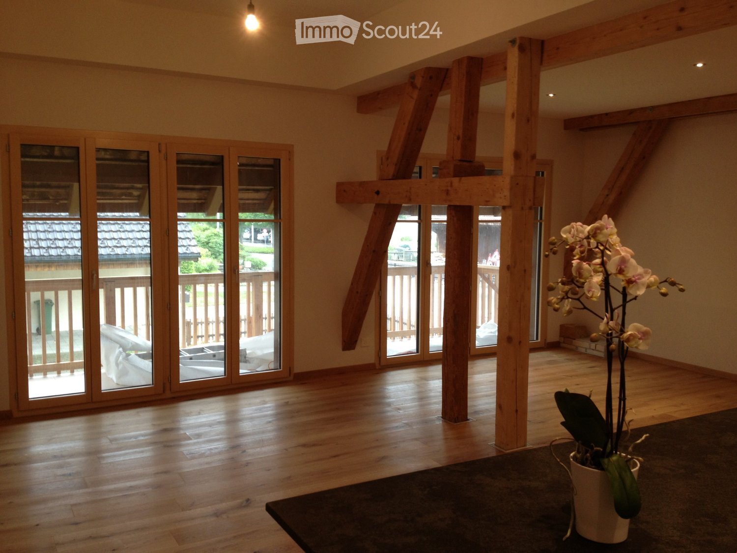 Bright, airy room with wooden beams, large glass doors leading to a balcony, wooden flooring, and a small table with a flower pot.